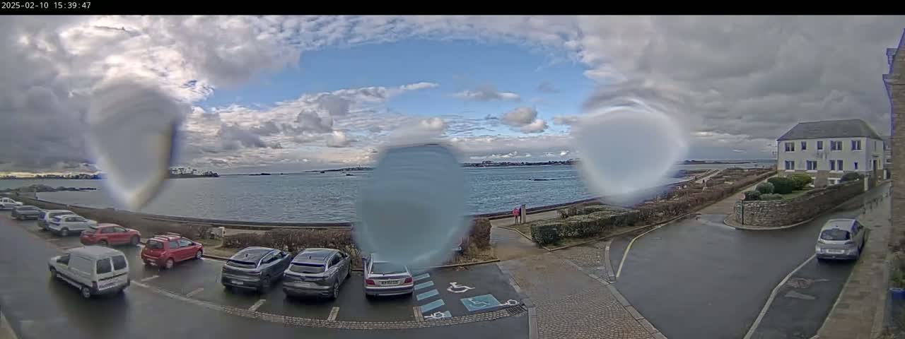 A partly cloudy day shows a waterfront view with several cars parked in a lot, a building, and a body of water beyond a walkway.