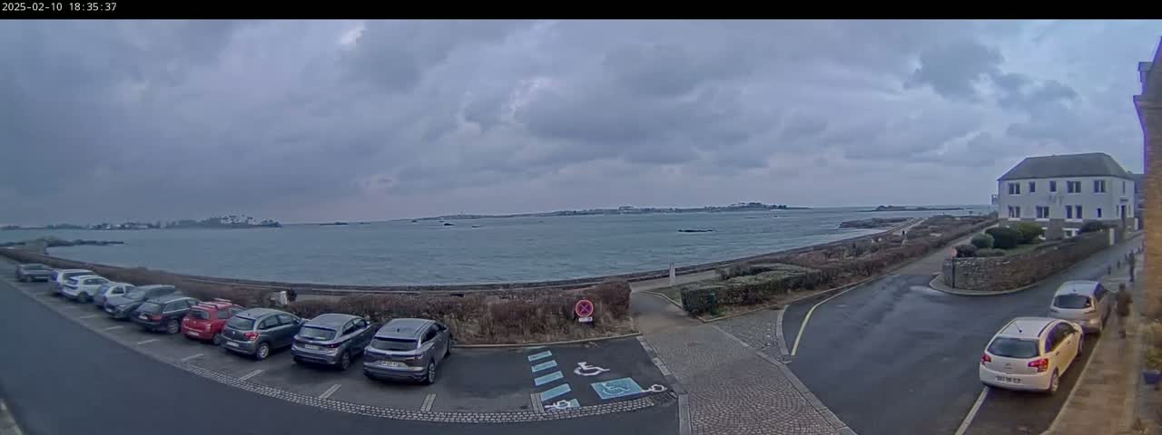 A parking lot with several cars is situated near a calm body of water under a cloudy sky, next to a building.