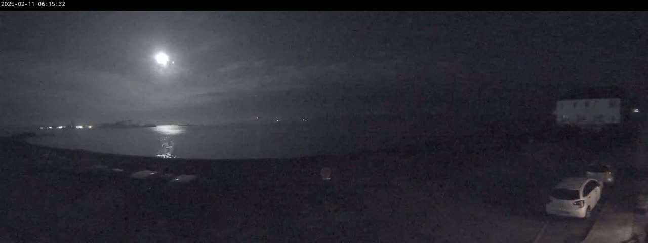 A nighttime view of a calm body of water under a bright moon, with a few cars parked in a lot nearby.