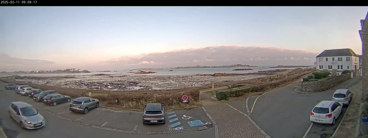 A parking lot with several cars is situated near a calm body of water under a partly cloudy, pale sky, with a distant shoreline and buildings visible.