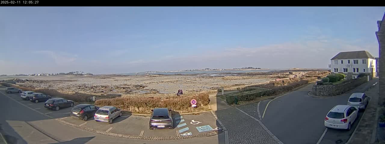 A mostly sunny day overlooks a coastal parking lot with several cars parked, a low tide revealing a rocky shoreline and distant land, and a two-story white building nearby.