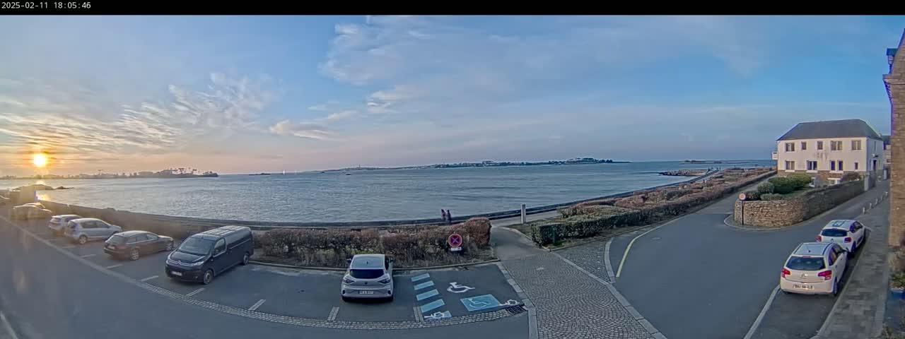 A seaside parking lot with several cars is seen at sunset under a mostly clear sky, overlooking a calm body of water and a distant coastline.