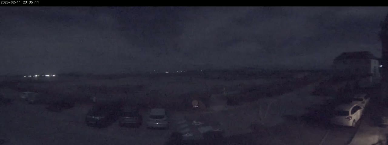 A nighttime view of a parking lot with several cars parked, under a dark, cloudy sky, with distant lights visible on the horizon.