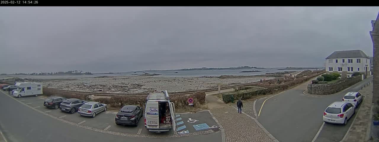 A parking lot with several cars and a van is situated near a rocky shoreline and calm ocean under an overcast sky.