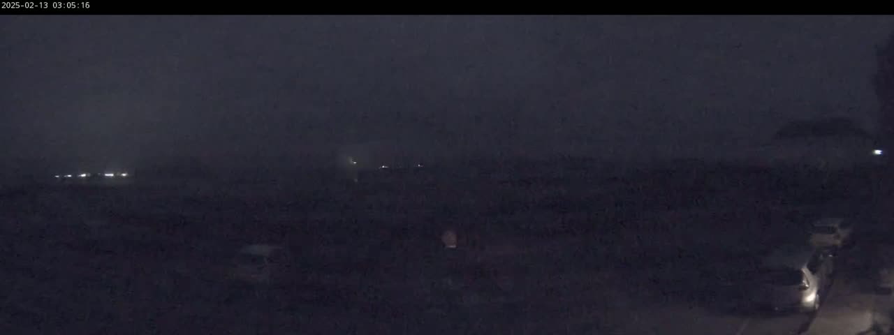 A dark, nighttime scene shows a few headlights in the distance and a few vehicles parked or driving closer to the camera under a hazy sky.