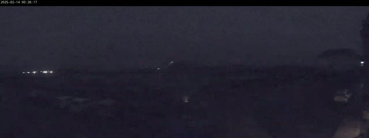 A dark, nighttime scene shows indistinct shapes and a few faint lights in the distance under a hazy sky.
