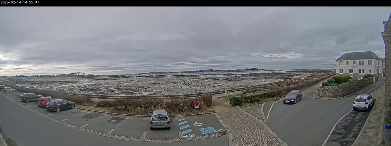 A parking lot with several cars is situated near a low stone wall overlooking a large expanse of flat, light-brown land extending to a calm, grey-toned sea under an overcast sky.