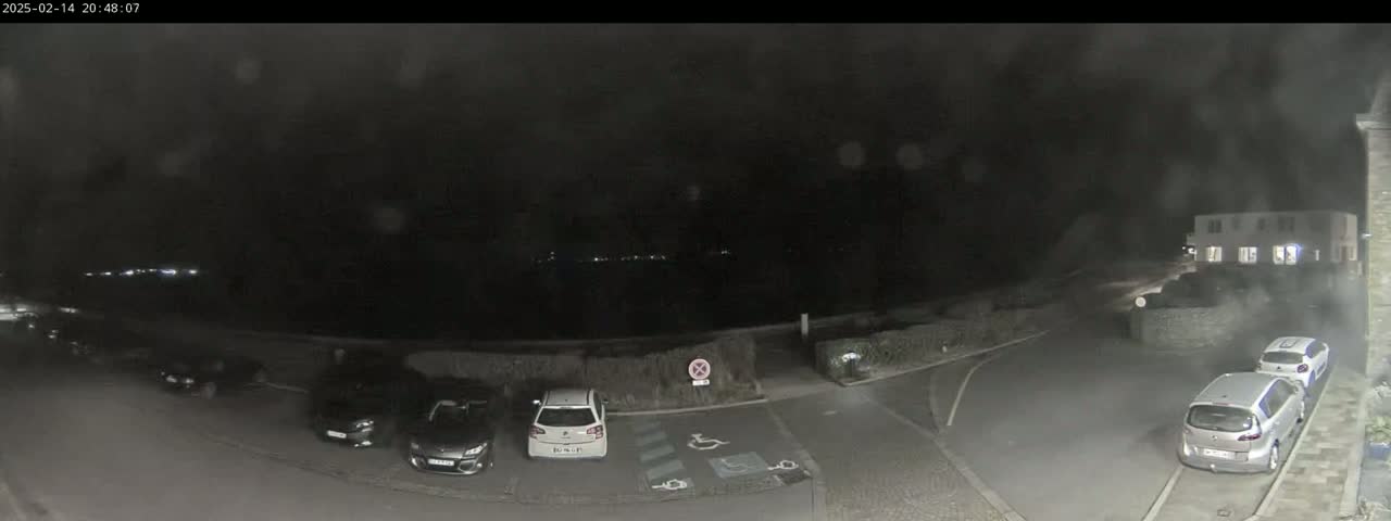 A nighttime, rain-streaked view of a parking lot with several cars parked, and a building in the background.