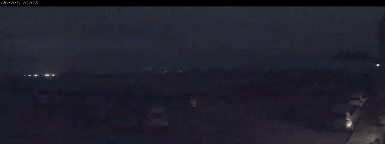 A dark, nighttime scene shows a few cars parked in a lot with dimly lit headlights visible in the distance.