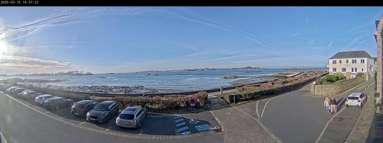 A seaside parking lot with several cars is next to a calm ocean under a mostly clear blue sky.