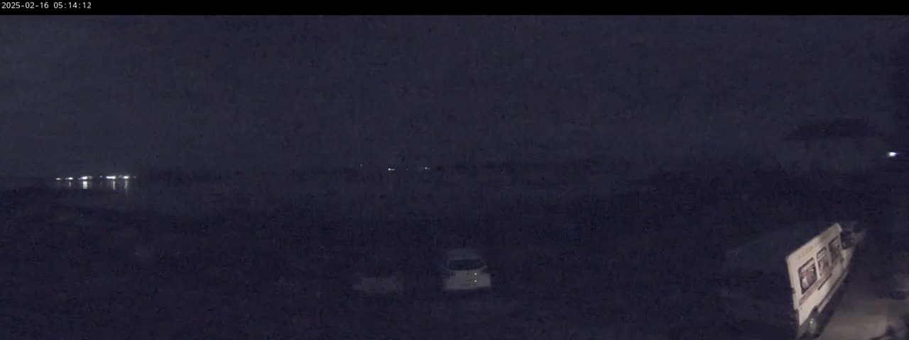 A nighttime, dark scene shows a few vehicles parked in a lot with dim lights visible in the distance across water.