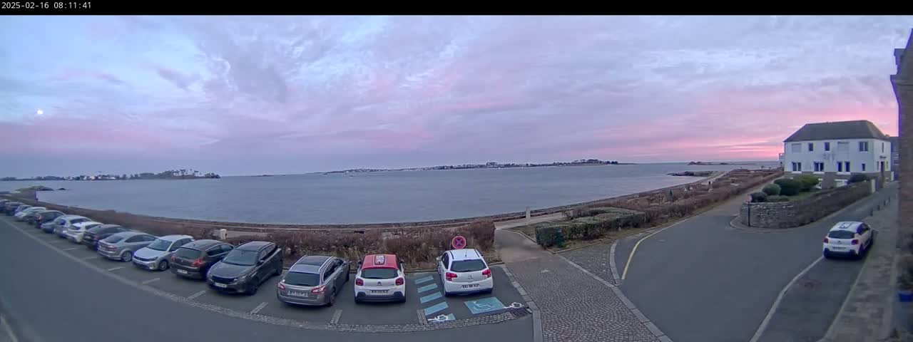 A parking lot with several cars is situated next to a calm body of water under a pale pink and purple sunrise sky.