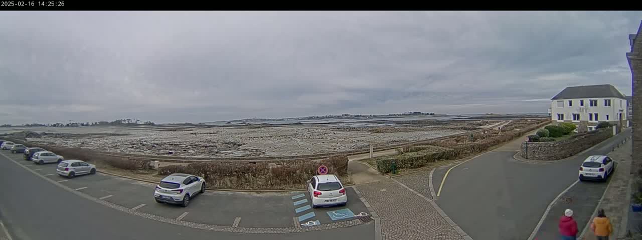 A parking lot with several cars is situated near a coastal area with rocky terrain under an overcast sky.