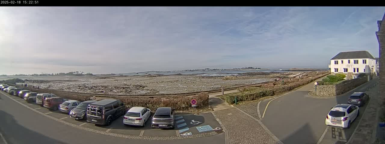 A parking lot with several cars is situated near a coastal area with low tide, under a mostly clear sky.