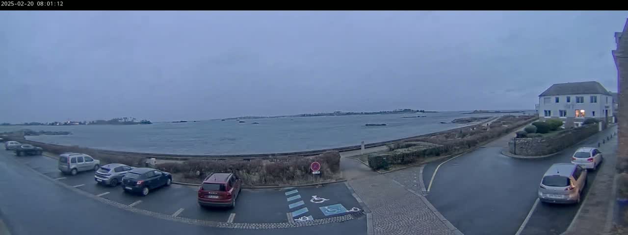A calm, overcast day reveals a seaside parking lot with several cars parked near a building and a body of water with a distant shoreline.