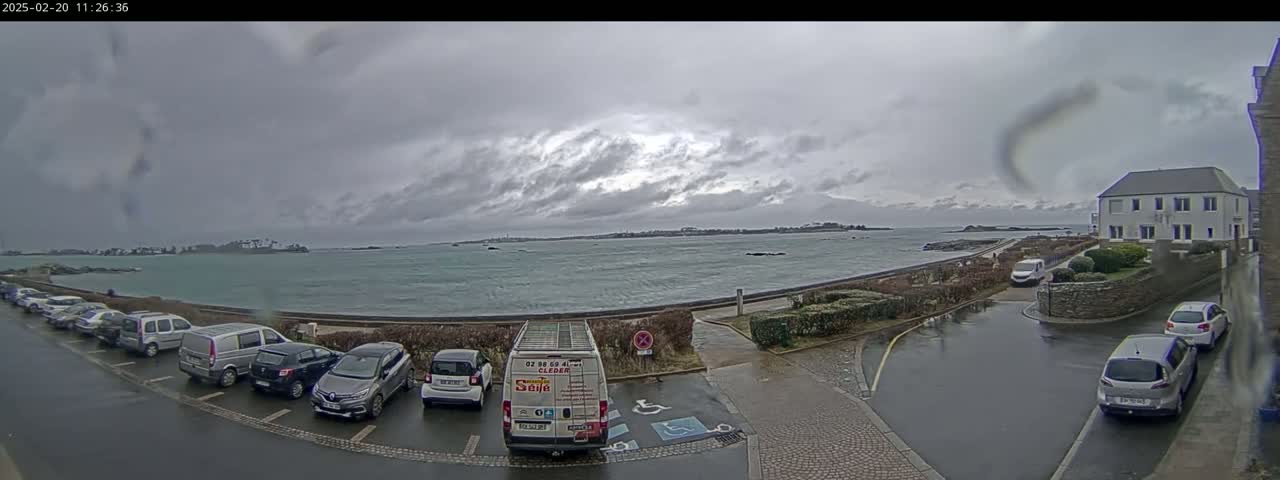 A parking lot with several cars is next to a waterfront road with a choppy sea under an overcast sky.