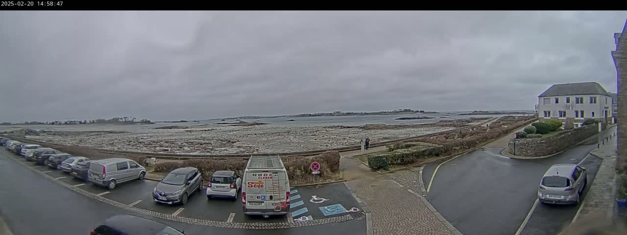 A parking lot with several cars and a van is situated near a coastal road, overlooking a calm body of water under an overcast sky.