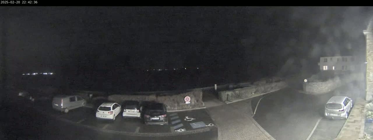 A nighttime, hazy scene shows several cars parked in a lot adjacent to a stone wall and building.