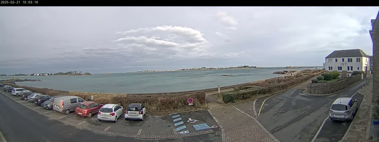 A partly cloudy day overlooks a calm body of water with cars parked in a lot near a building and a shoreline.