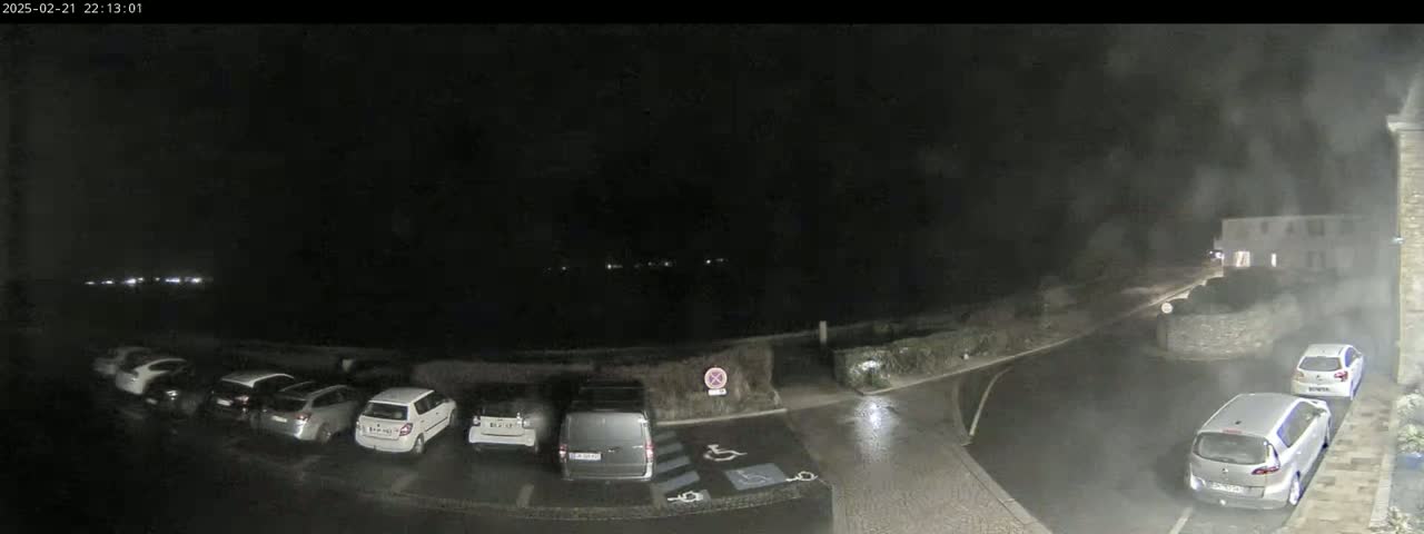 A nighttime, wet parking lot with several cars parked, near a building and a road.