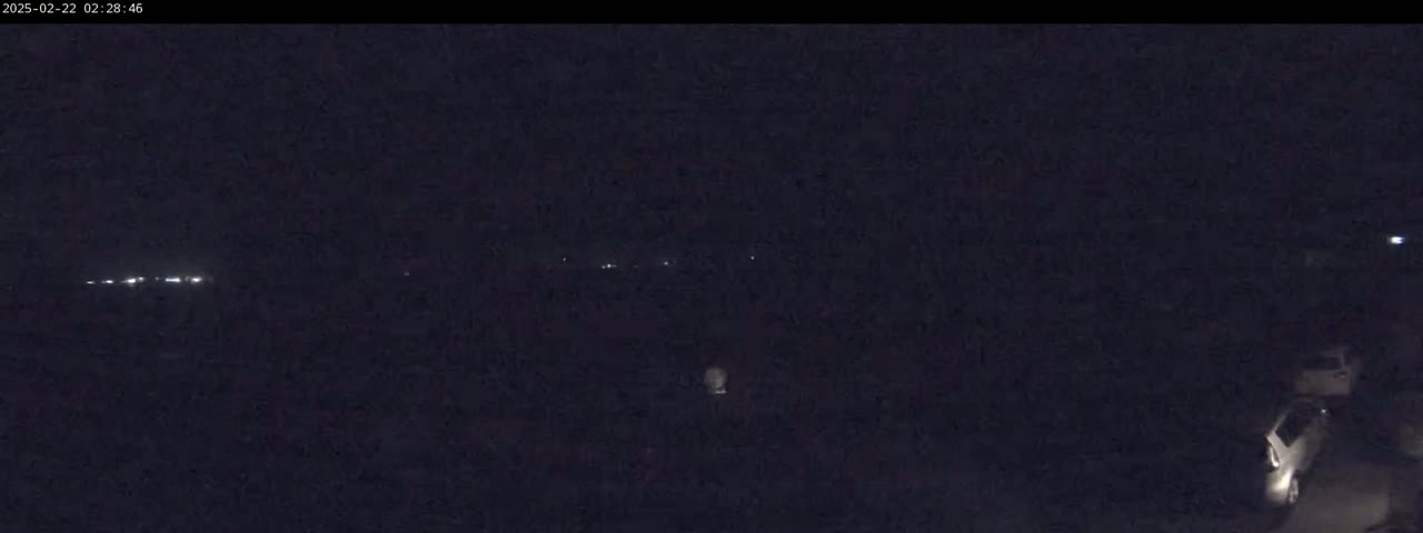 A nighttime, low-light view shows a dark landscape with a few distant lights and a vehicle in the lower right corner.
