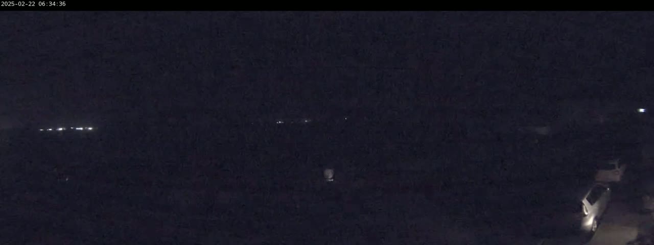A nighttime, low-light view shows a dark, mostly featureless landscape with a few distant lights and a couple of vehicles in the lower right corner.