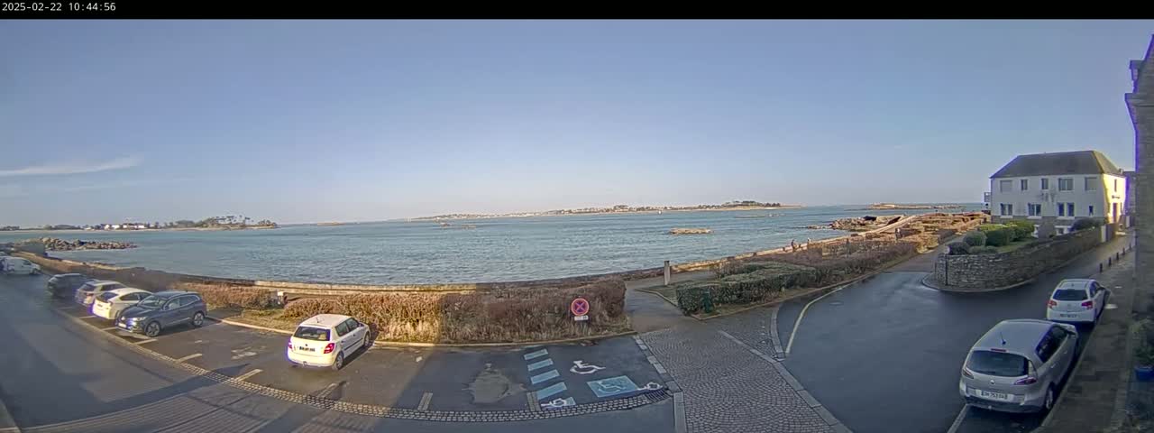 A clear, sunny day overlooks a calm body of water with a small town and parked cars visible near a stone wall.