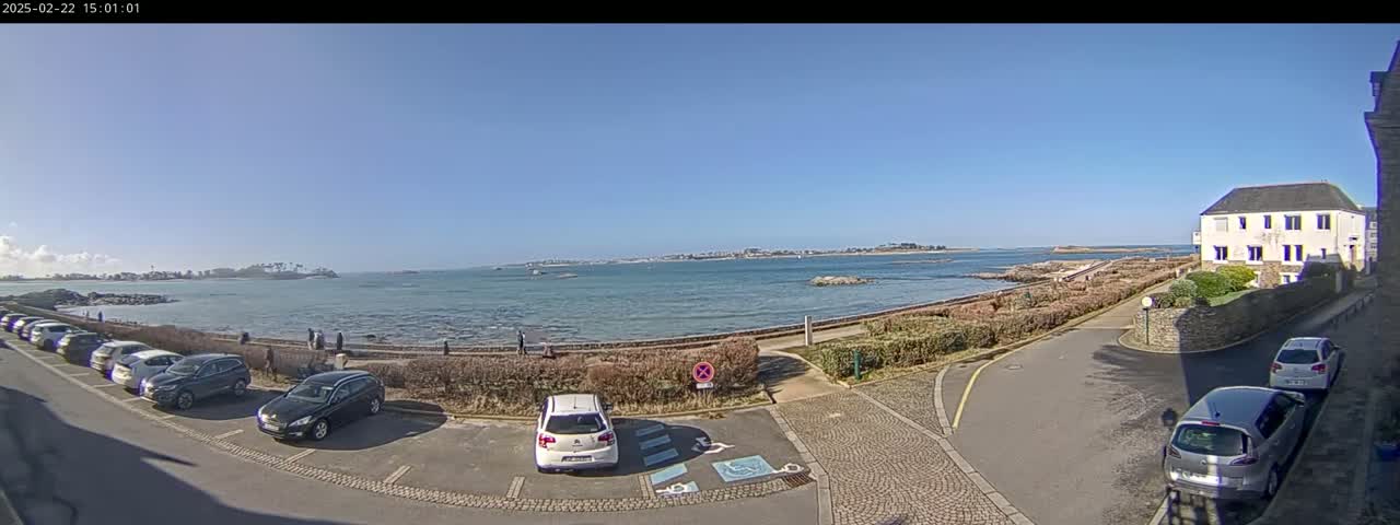 A seaside parking lot with several cars is next to a low stone wall overlooking calm water and distant land under a clear blue sky.