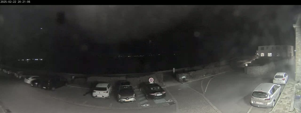 A nighttime, dimly lit parking lot with several cars parked, overlooks a dark body of water under a cloudy sky.