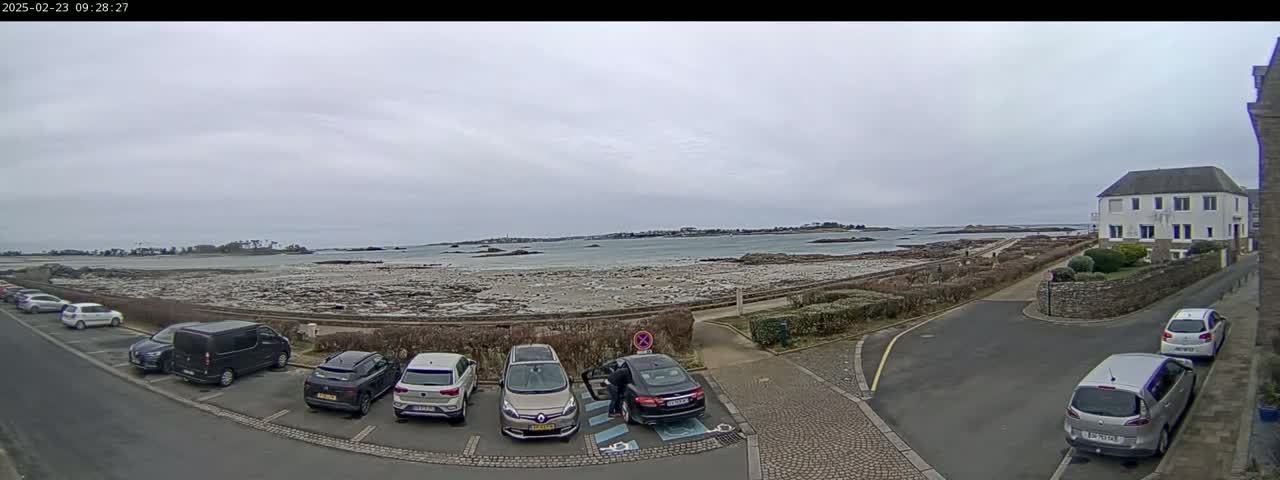 A parking lot with several cars is situated near a coastline under an overcast sky, overlooking a calm body of water and a small island in the distance.