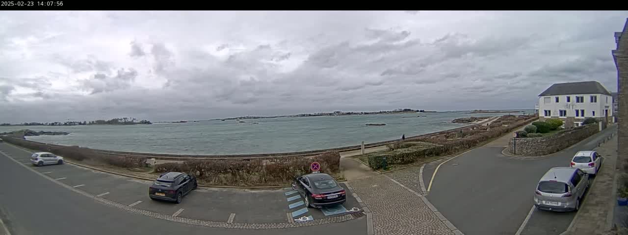 A coastal parking lot with several cars, overlooking a calm body of water under a cloudy sky.
