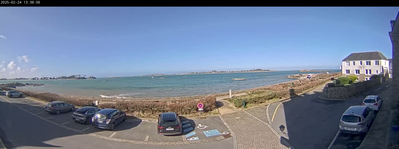 A sunny day shows a coastal scene with several cars parked near a building overlooking a calm body of water and distant land.