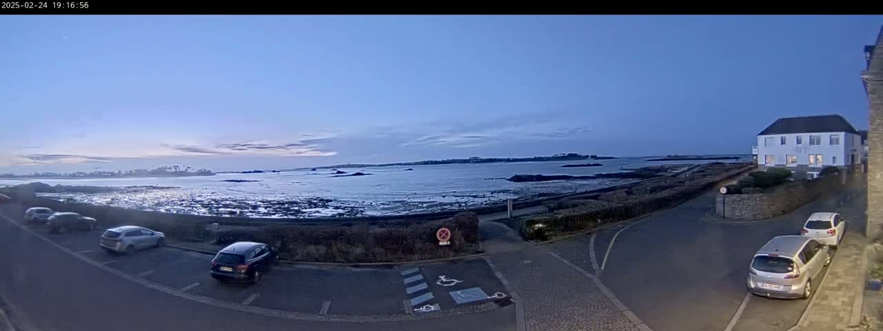 A seaside parking lot with several cars is visible, overlooking a calm body of water under a twilight sky.