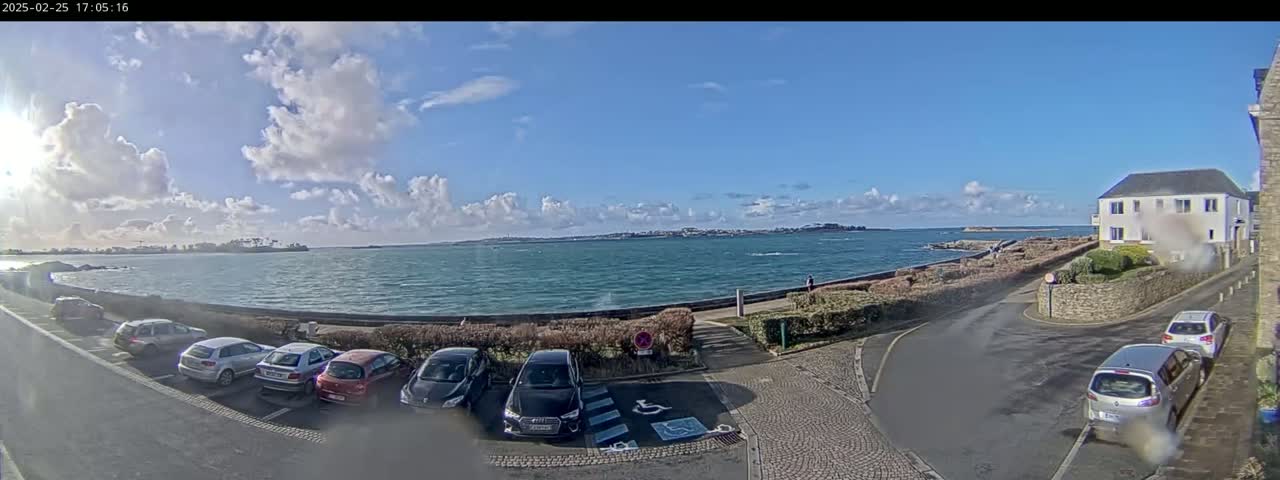 A partly sunny day shows a coastal scene with a body of water, several parked cars, and a building, all viewed from an elevated position.