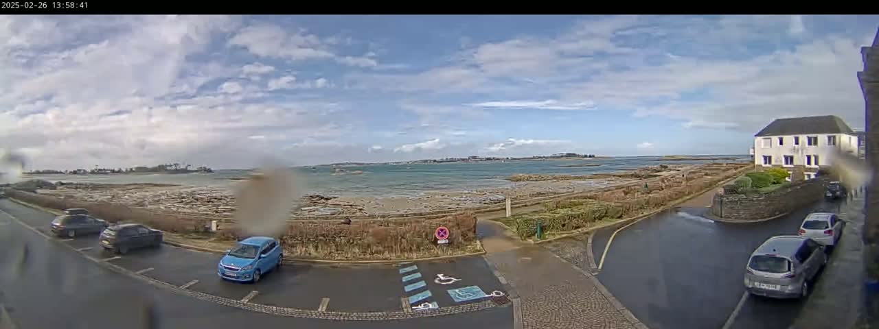 A partly cloudy day reveals a coastal scene with a few cars parked near a building and a body of water with a shoreline of rocks and low vegetation.