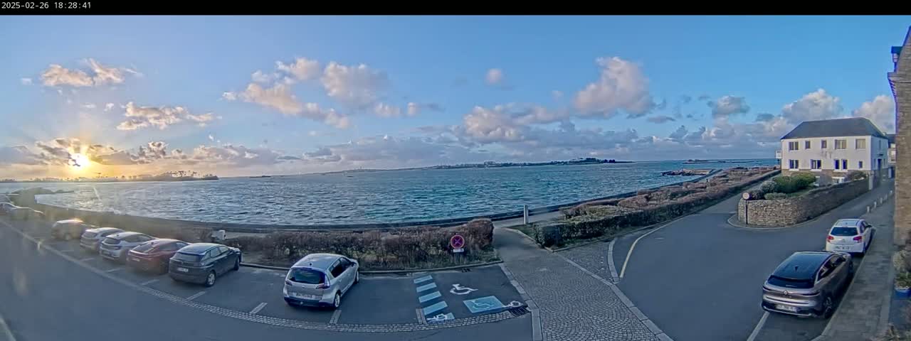 A seaside parking lot with several cars parked, overlooking a wavy body of water under a partly cloudy sunset sky.