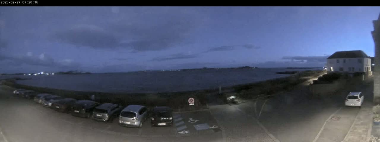 A parking lot with several cars is situated near a calm body of water under a twilight sky with some clouds.