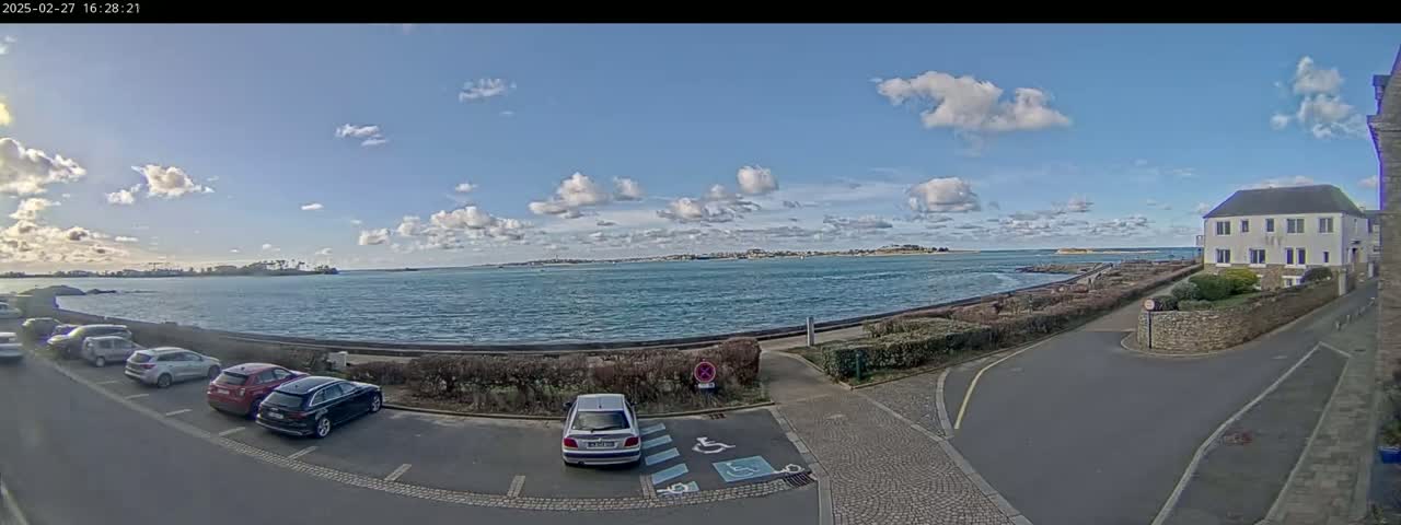 Several cars are parked in a lot overlooking a calm body of water under a partly cloudy sky.