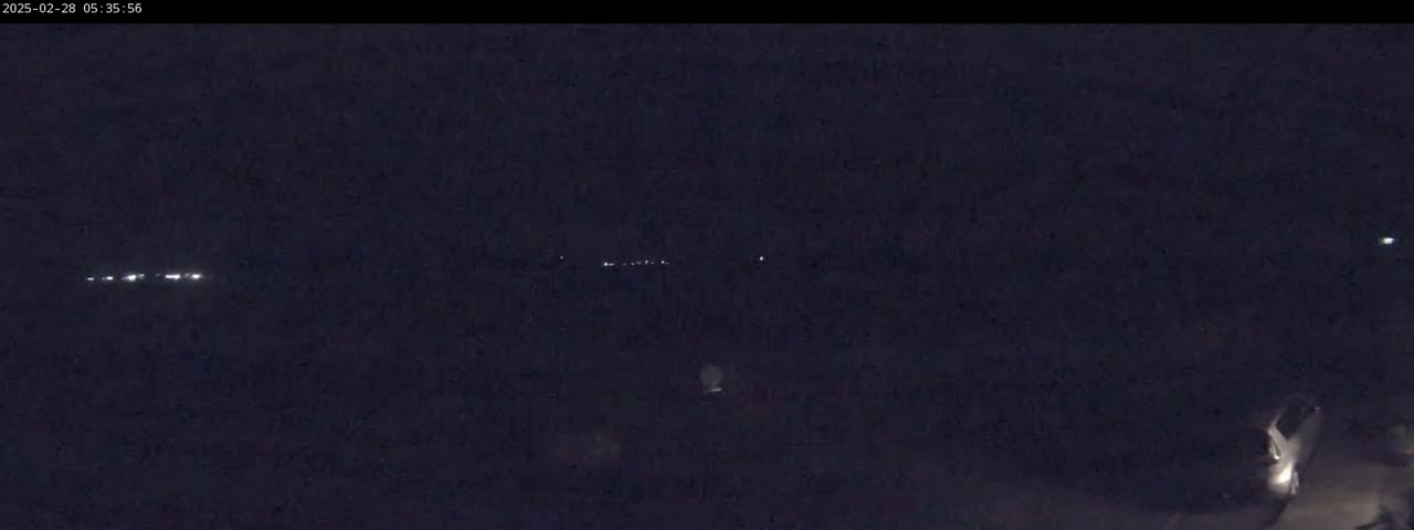 A dark, nighttime scene shows distant lights in the background and a vehicle parked in the lower right corner.