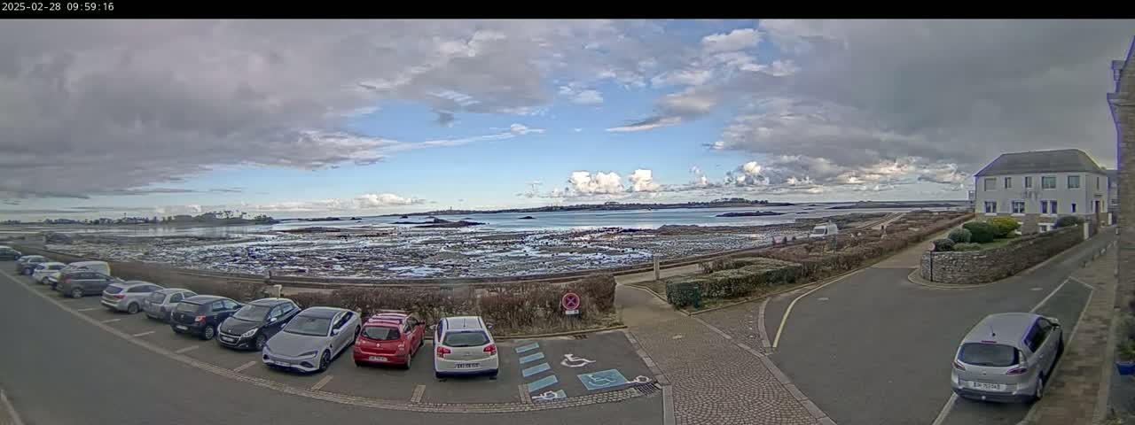 A partly cloudy day reveals a coastal scene with a parking lot full of cars near a body of water and a low, light-colored house.