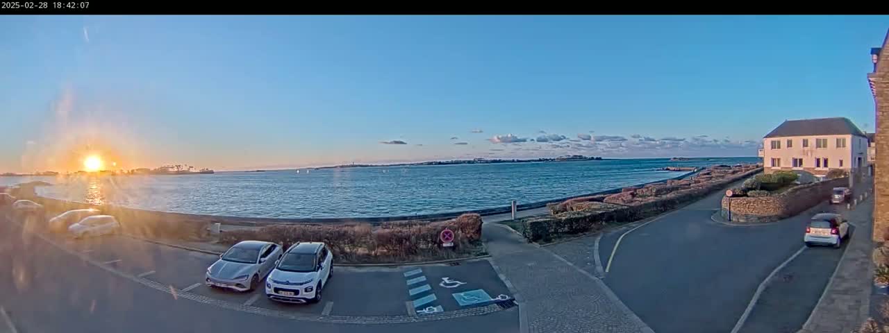 A seaside parking lot with several cars, a calm body of water reflecting the setting sun under a mostly clear sky, and a building in the background.