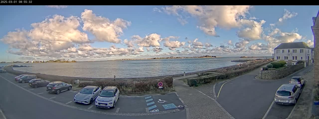 A partly cloudy day overlooks a calm body of water with cars parked in a lot next to a building.