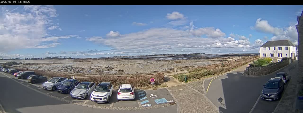 A partly cloudy day reveals a coastal scene with several cars parked near a building and a wide expanse of exposed tidal flats stretching to a distant shoreline.