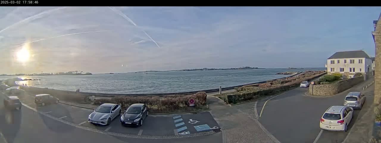 A partly sunny day shows a calm body of water with a distant shoreline, several parked cars in a lot near a building, and a roadway leading to the water's edge.