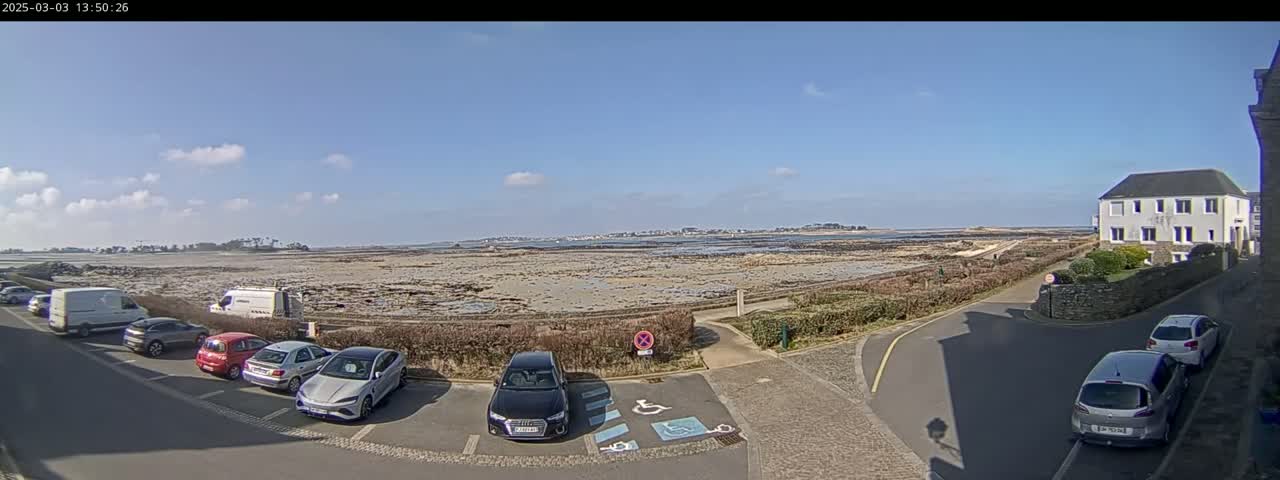 A seaside parking lot with several cars parked, overlooks a flat expanse of exposed land and a calm body of water under a mostly clear, sunny sky.