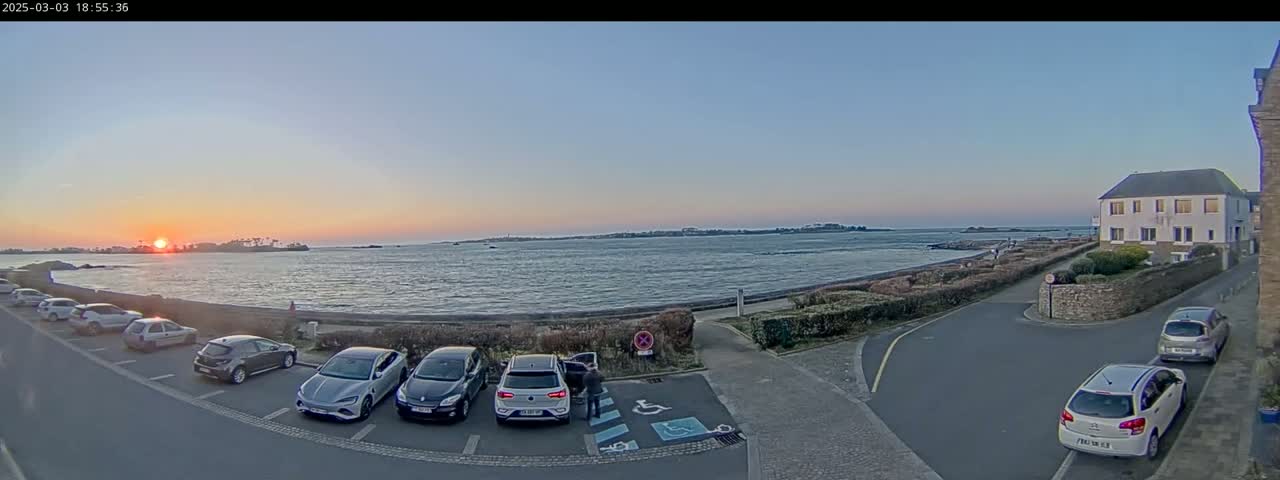 A sunset over a calm body of water is visible from a parking lot next to a road and a building, with several cars parked nearby.