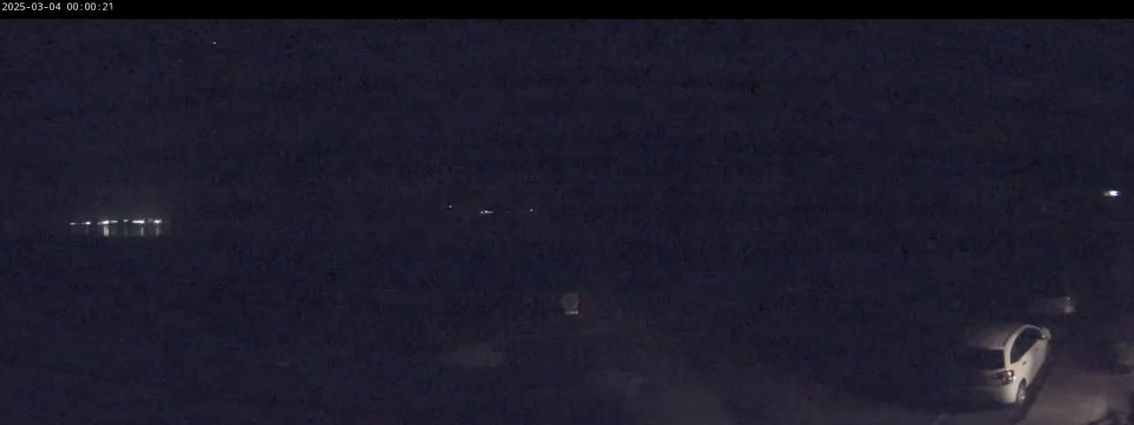 A dark, nighttime scene shows a body of water with faint lights in the distance and a white car parked in a dark area closer to the camera.