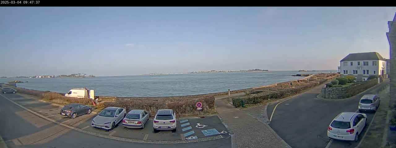 A clear, sunny day overlooking a calm body of water with several cars parked near a building and a stone wall.