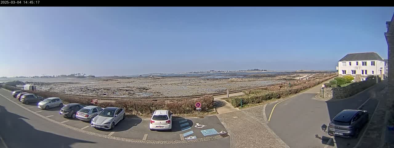A coastal parking lot with several cars is situated next to a building overlooking a rocky, low-tide beach and distant land on a sunny day.