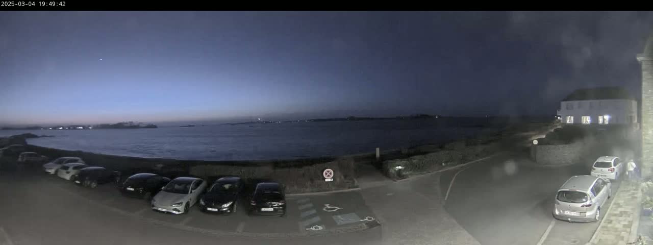 A twilight view shows a dark body of water, a parking lot with several cars, and a building, all under a clear, dark blue sky.
